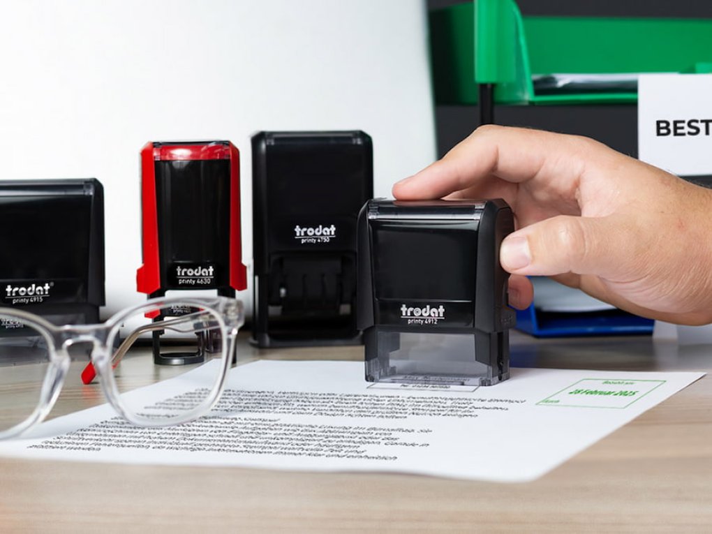 Hand greift nach einem Trodat-Stempel auf einem Schreibtisch mit Dokument und Brille